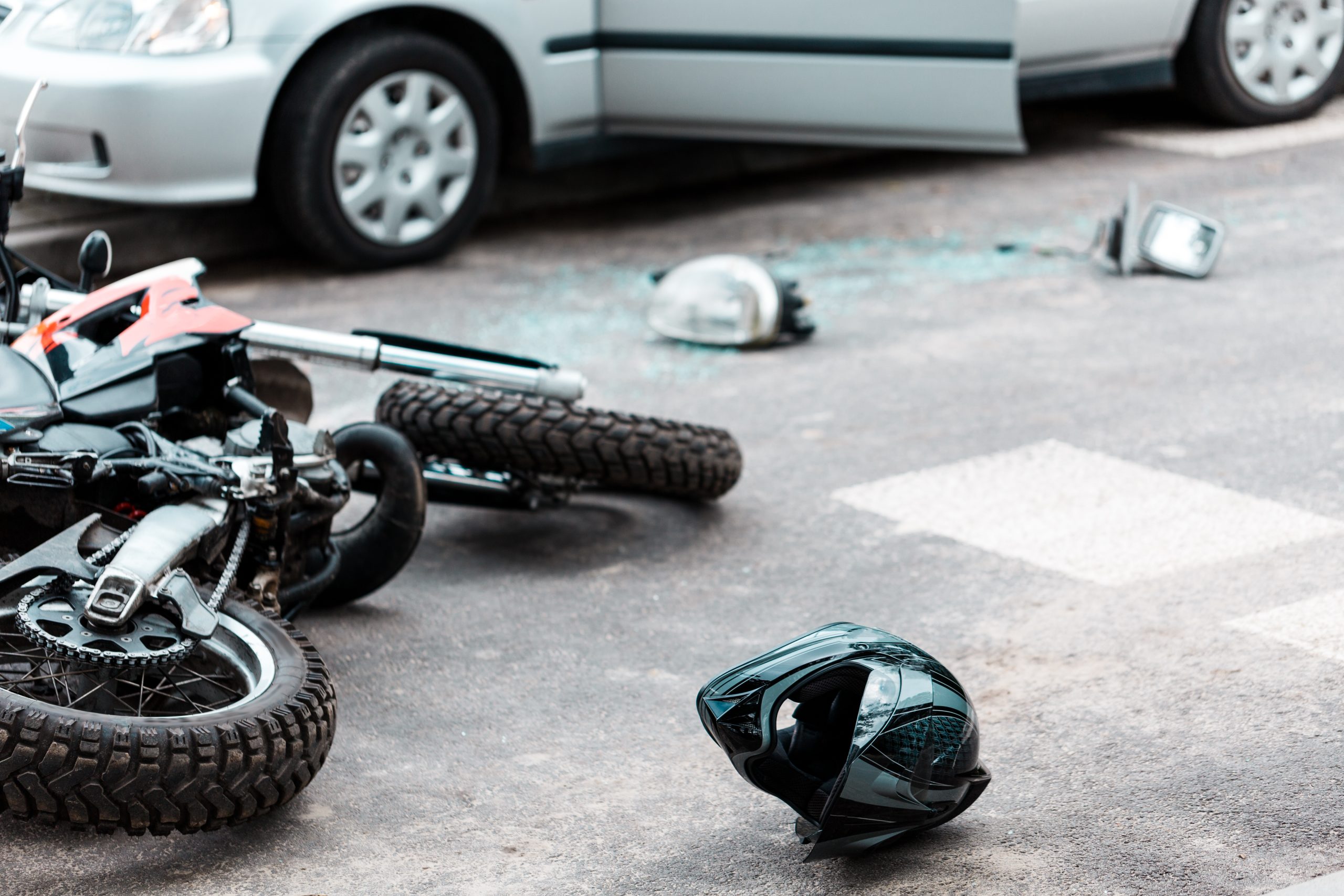Motorcycle lying on its side in the street beside a car after a collision, with a helmet and broken glass on the pavement
