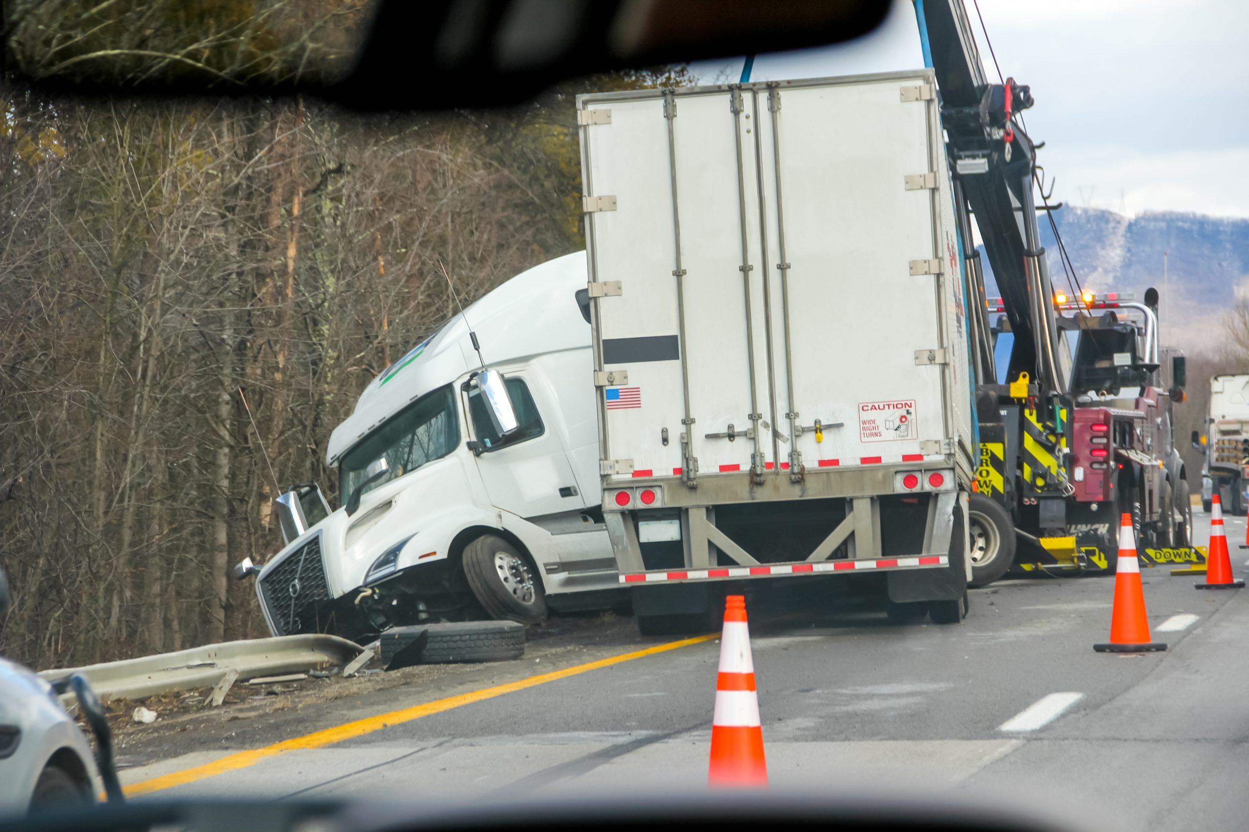 Semi-truck with a box trailer run off a highway into the guardrail, with a tow recovery unit and traffic cones at the scene